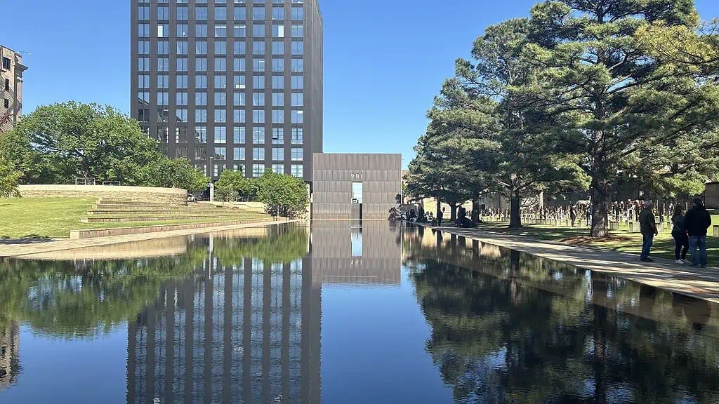 Reflecting pool in a city park with a tall dark office building on the left, trees on the right, and people strolling along the walkway.