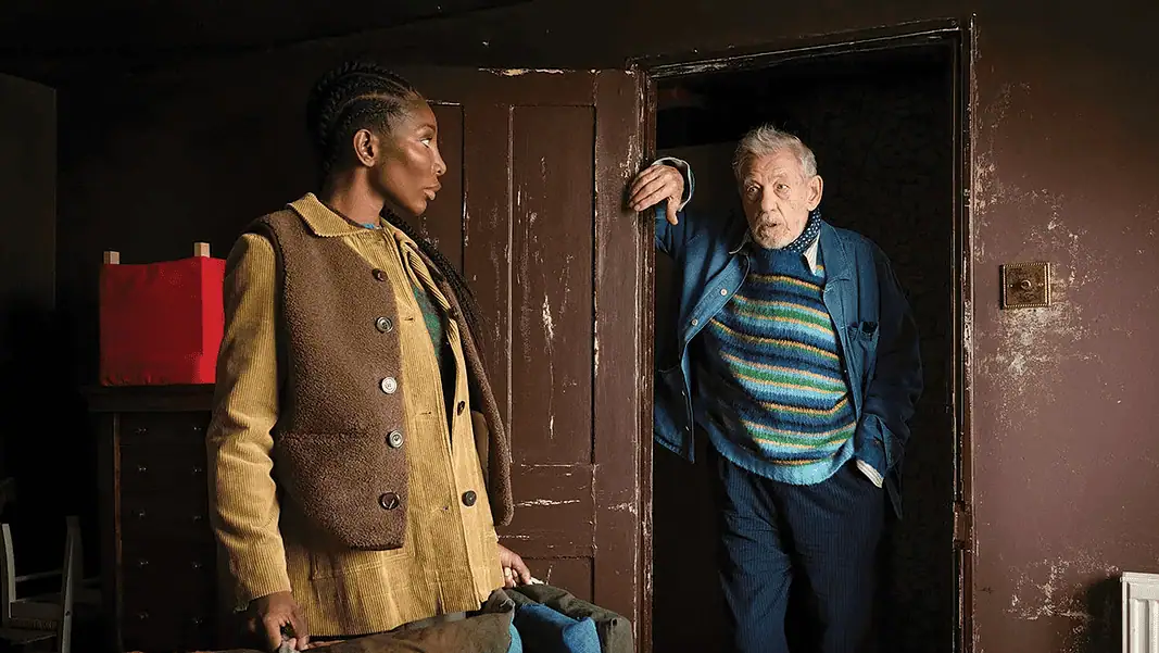 The Christophers 1 A young woman in a tan corduroy jacket stands in a room while an older man in a striped sweater steps through a dark doorway, gesturing with one hand.