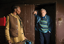 A young woman in a tan corduroy jacket stands in a room while an older man in a striped sweater steps through a dark doorway, gesturing with one hand.