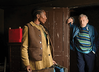 A young woman in a tan corduroy jacket stands in a room while an older man in a striped sweater steps through a dark doorway, gesturing with one hand.