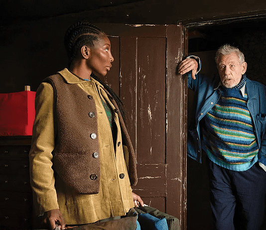 A young woman in a tan corduroy jacket stands in a room while an older man in a striped sweater steps through a dark doorway, gesturing with one hand.