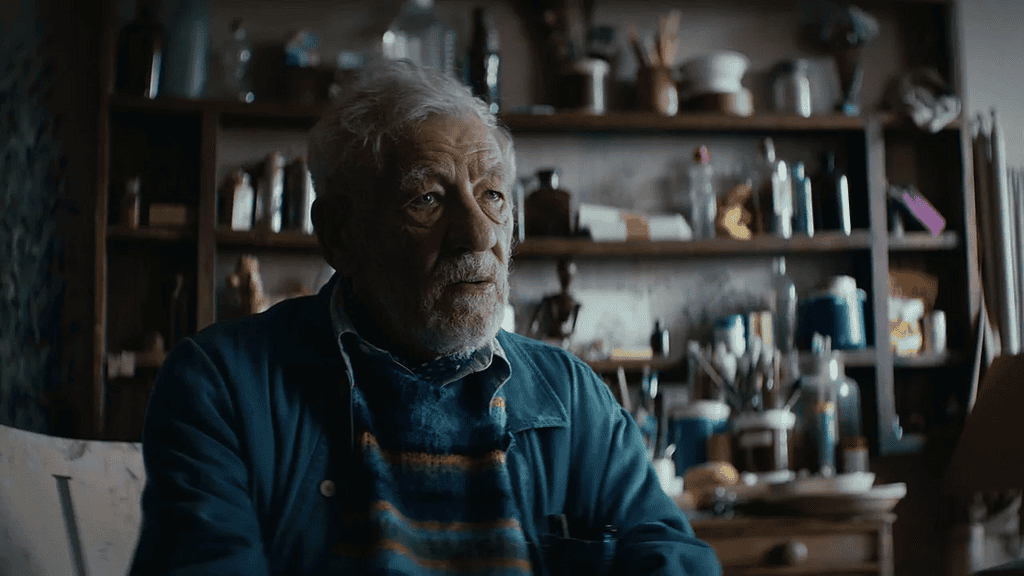 A thoughtful elderly man with gray hair and beard sits in a cluttered workshop, looking off to the side.