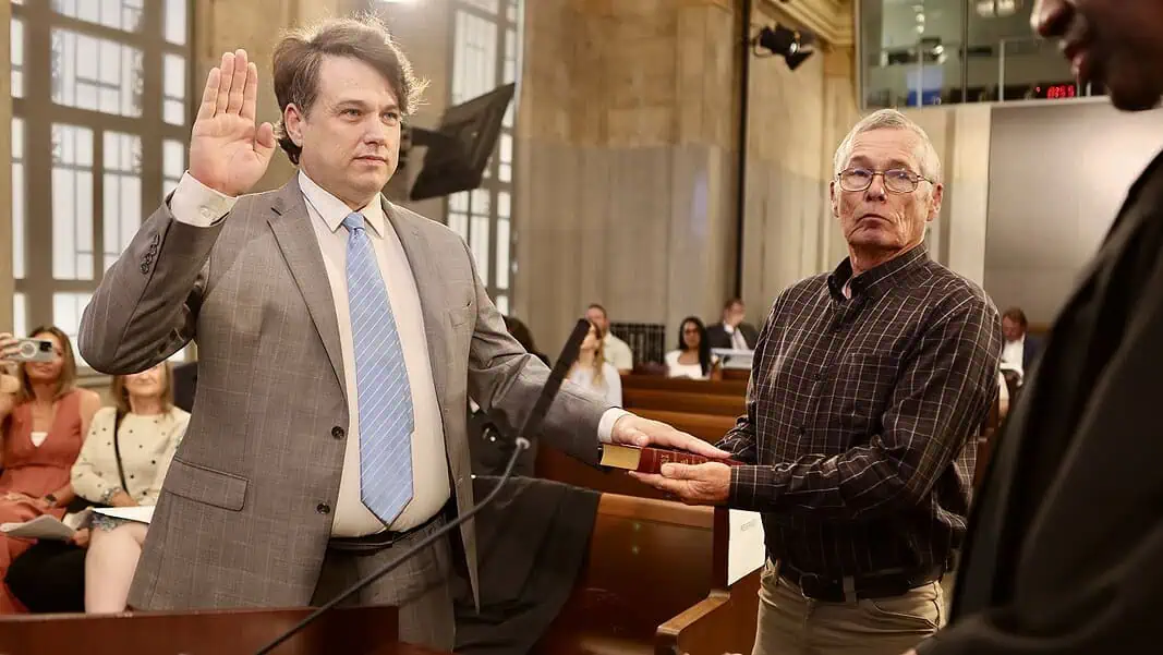 judge-young-swearing-in Man in a gray suit raises his right hand to take an oath, as another man holds a book for him in a formal setting.