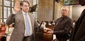 Man in a gray suit raises his right hand to take an oath, as another man holds a book for him in a formal setting.