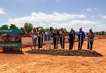 Groundbreaking ceremony for the Tom and Judy Love YMCA with people in hard hats holding shovels beside the sign.