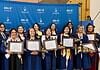 Group of graduates in blue gowns smiling and holding framed diplomas in front of a blue backdrop with OGE logos behind them.