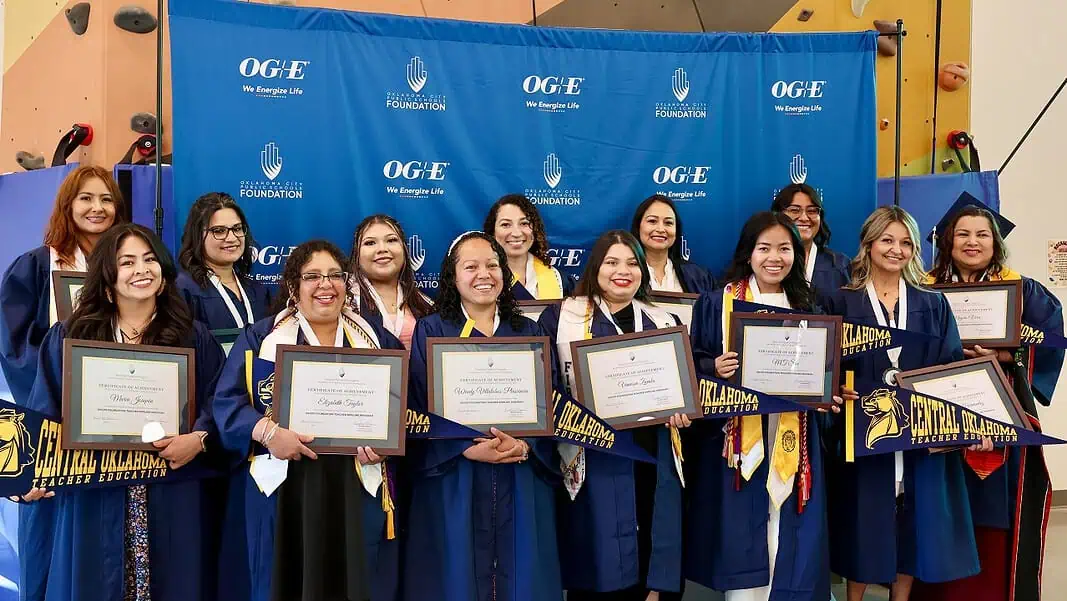 1S3A7174 Group of graduates in blue gowns smiling and holding framed diplomas in front of a blue backdrop with OGE logos behind them.