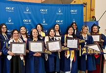 Group of graduates in blue gowns smiling and holding framed diplomas in front of a blue backdrop with OGE logos behind them.