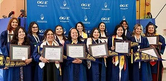 Group of graduates in blue gowns smiling and holding framed diplomas in front of a blue backdrop with OGE logos behind them.