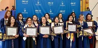 Group of graduates in blue gowns smiling and holding framed diplomas in front of a blue backdrop with OGE logos behind them.