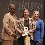 PRESS RELEASE: Karlie Minson named 2026 OKCPS District Teacher of the Year Four women in business attire on a stage; one woman in leopard print holds an award and a gift bag, flanked by three others celebrating.
