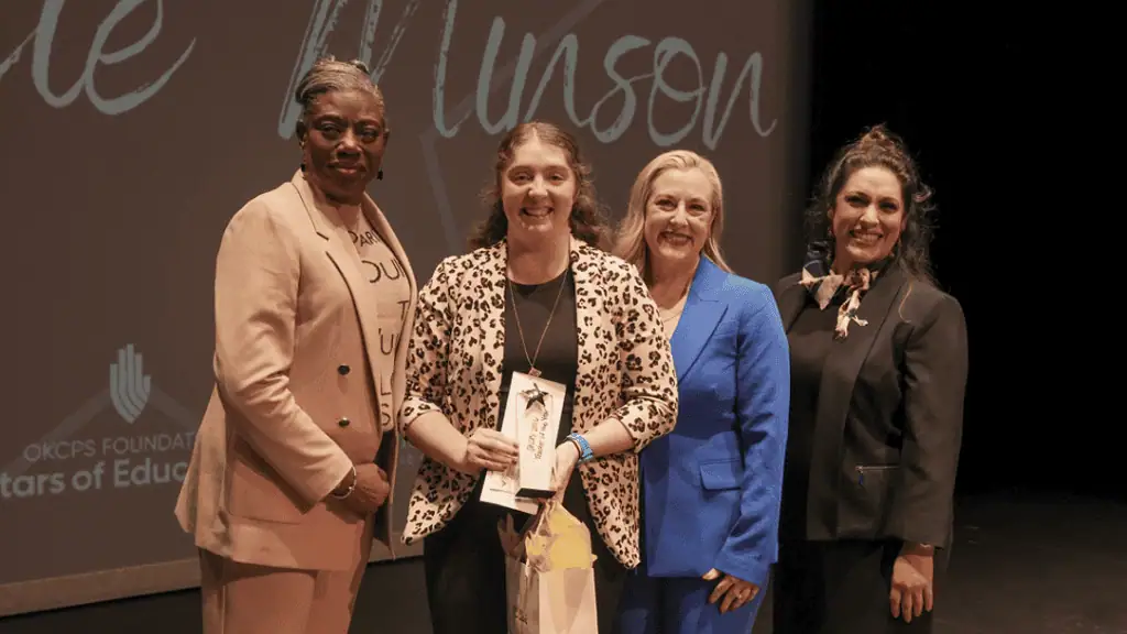 2026 OKCPS Teacher of the Year Minson Four women in business attire on a stage; one woman in leopard print holds an award and a gift bag, flanked by three others celebrating.