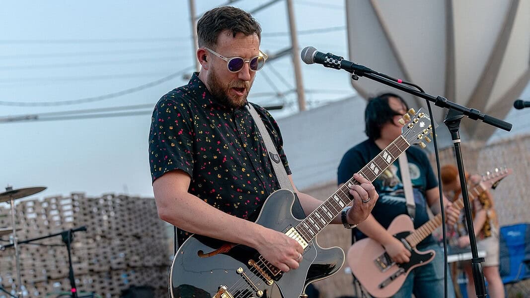 Male guitarist wearing sunglasses, playing a shiny gray electric guitar on an outdoor stage with a microphone in front of him.
