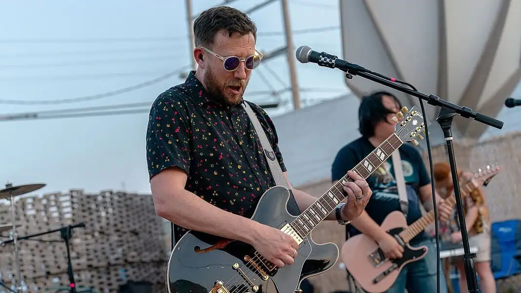 Fair Weather Enemies Male guitarist wearing sunglasses, playing a shiny gray electric guitar on an outdoor stage with a microphone in front of him.
