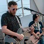 Rock out without your wallet out with these free concerts in May Male guitarist wearing sunglasses, playing a shiny gray electric guitar on an outdoor stage with a microphone in front of him.