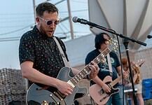 Male guitarist wearing sunglasses, playing a shiny gray electric guitar on an outdoor stage with a microphone in front of him.