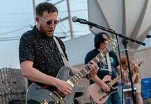 Rock out without your wallet out with these free concerts in May Male guitarist wearing sunglasses, playing a shiny gray electric guitar on an outdoor stage with a microphone in front of him.
