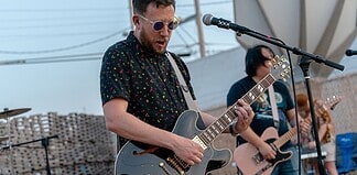 Male guitarist wearing sunglasses, playing a shiny gray electric guitar on an outdoor stage with a microphone in front of him.