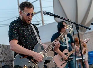 Rock out without your wallet out with these free concerts in May Male guitarist wearing sunglasses, playing a shiny gray electric guitar on an outdoor stage with a microphone in front of him.