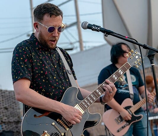 Male guitarist wearing sunglasses, playing a shiny gray electric guitar on an outdoor stage with a microphone in front of him.
