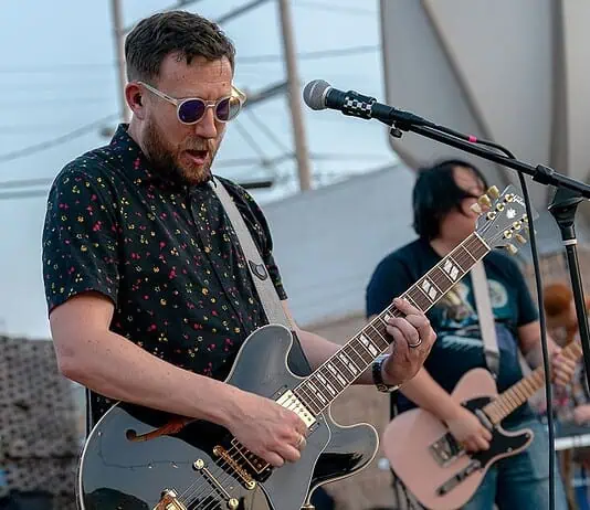 Rock out without your wallet out with these free concerts in May Male guitarist wearing sunglasses, playing a shiny gray electric guitar on an outdoor stage with a microphone in front of him.