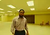 May movies explore mystery, madness, meditation of liminal space A man in a beige button-down shirt stands in a large empty room with yellow lighting and scattered wooden chairs in the background.