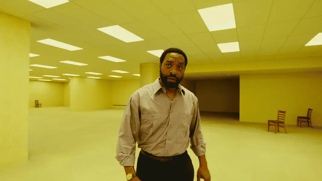 Backrooms 2 A man in a beige button-down shirt stands in a large empty room with yellow lighting and scattered wooden chairs in the background.