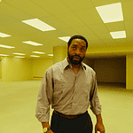 A man in a beige button-down shirt stands in a large empty room with yellow lighting and scattered wooden chairs in the background.