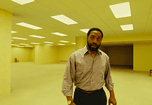 A man in a beige button-down shirt stands in a large empty room with yellow lighting and scattered wooden chairs in the background.
