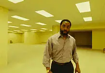May movies explore mystery, madness, meditation of liminal space A man in a beige button-down shirt stands in a large empty room with yellow lighting and scattered wooden chairs in the background.