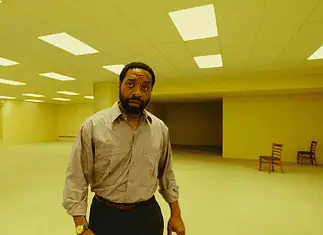 May movies explore mystery, madness, meditation of liminal space A man in a beige button-down shirt stands in a large empty room with yellow lighting and scattered wooden chairs in the background.