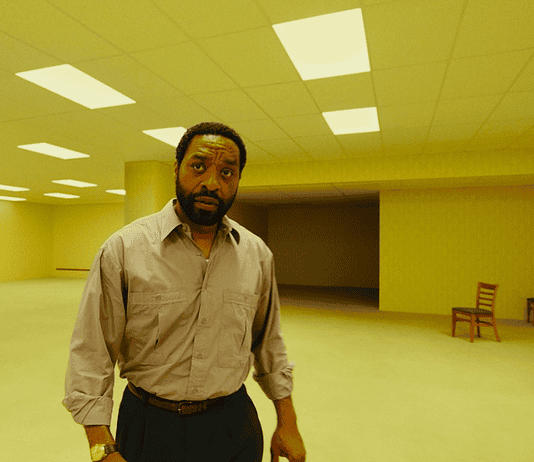 A man in a beige button-down shirt stands in a large empty room with yellow lighting and scattered wooden chairs in the background.