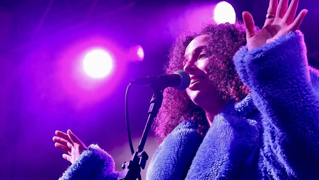 Female singer singing into a microphone on a concert stage under purple lights, wearing a blue fluffy coat