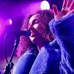 Love is in the spring air with songs of intensity, obsession, regret Female singer singing into a microphone on a concert stage under purple lights, wearing a blue fluffy coat