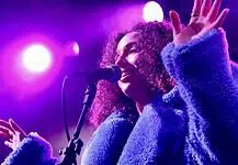 Love is in the spring air with songs of intensity, obsession, regret Female singer singing into a microphone on a concert stage under purple lights, wearing a blue fluffy coat