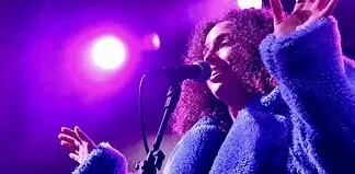 Female singer singing into a microphone on a concert stage under purple lights, wearing a blue fluffy coat