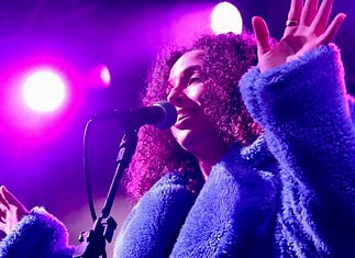Female singer singing into a microphone on a concert stage under purple lights, wearing a blue fluffy coat