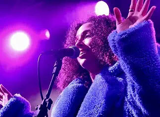 Love is in the spring air with songs of intensity, obsession, regret Female singer singing into a microphone on a concert stage under purple lights, wearing a blue fluffy coat