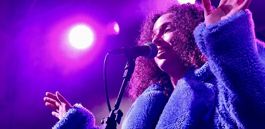 Female singer singing into a microphone on a concert stage under purple lights, wearing a blue fluffy coat