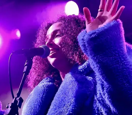Love is in the spring air with songs of intensity, obsession, regret Female singer singing into a microphone on a concert stage under purple lights, wearing a blue fluffy coat