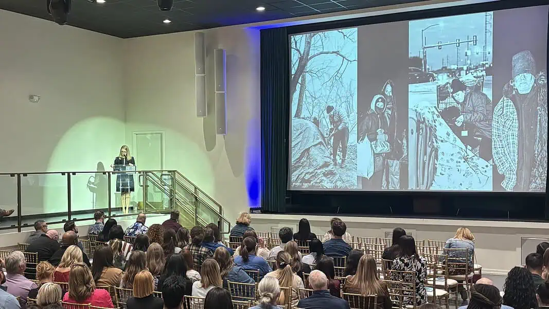 Caves homelessness 4 Woman in black dress speaking at a clear podium on a stage, audience seated in rows in a large auditorium, with a large grayscale collage projected on a screen to the right