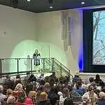 OKC reports first drop in homelessness in years, but challenges remain Woman in black dress speaking at a clear podium on a stage, audience seated in rows in a large auditorium, with a large grayscale collage projected on a screen to the right