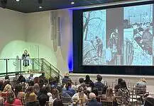 OKC reports first drop in homelessness in years, but challenges remain Woman in black dress speaking at a clear podium on a stage, audience seated in rows in a large auditorium, with a large grayscale collage projected on a screen to the right