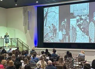 OKC reports first drop in homelessness in years, but challenges remain Woman in black dress speaking at a clear podium on a stage, audience seated in rows in a large auditorium, with a large grayscale collage projected on a screen to the right