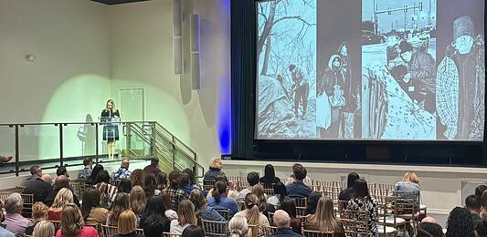 Woman in black dress speaking at a clear podium on a stage, audience seated in rows in a large auditorium, with a large grayscale collage projected on a screen to the right