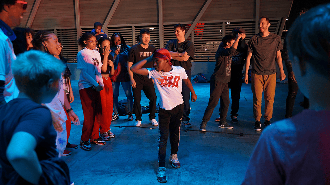 Group of youths forming a circle around a dancer in a white shirt performing a breakdance move under blue lighting.