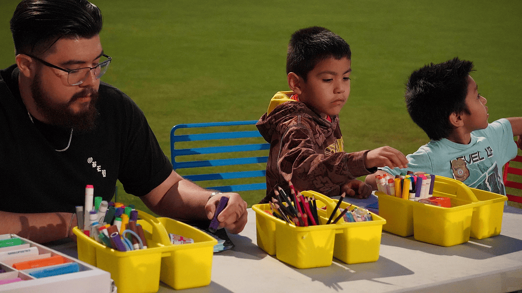 Man with glasses and beard sits at a table with yellow art bins while two young boys craft outdoors on a grassy field.