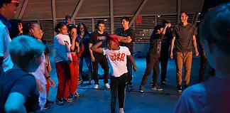 Group of youths forming a circle around a dancer in a white shirt performing a breakdance move under blue lighting.