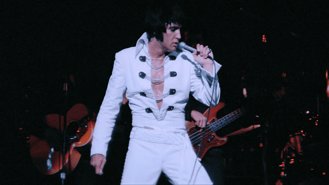 Elvis tribute artist in a white jumpsuit performs on stage, singing into a microphone with a guitarist in the background.