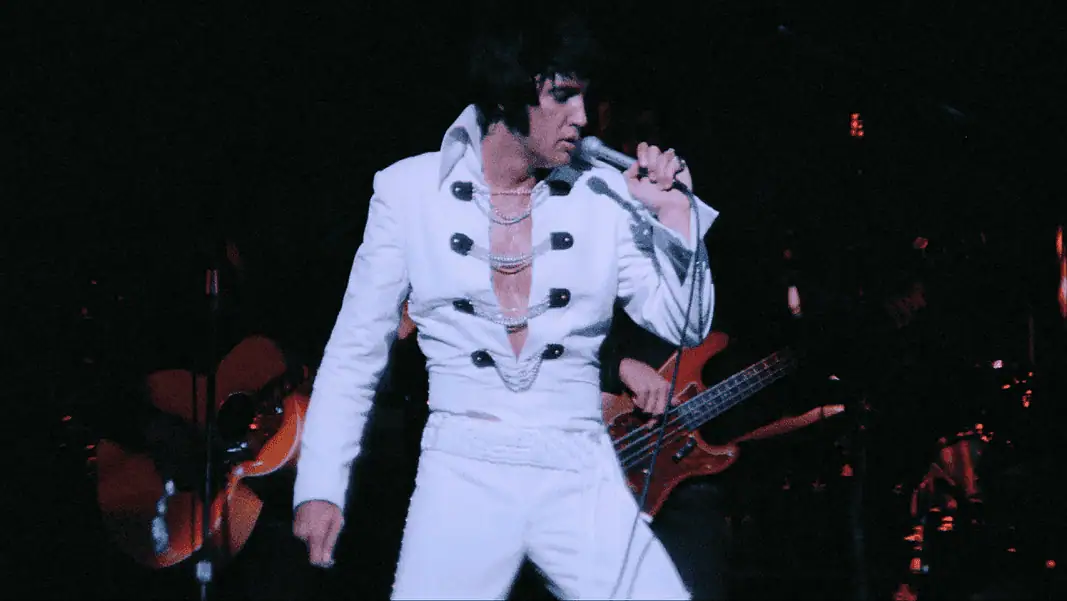 EPiC 1 Elvis tribute artist in a white jumpsuit performs on stage, singing into a microphone with a guitarist in the background.